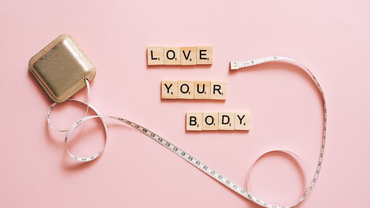 Scrabble letters spelling 'Love Your Body' with a tape measure on a pink backdrop.
